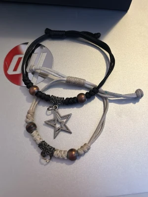 Två armband med stjärnhänge - Två justerbara armband i svart och beige snöre med dekorativa pärlor i metall och trä. Ena armbandet har ett hänge i form av en stjärna i silverfärgad metall. Perfekt för dig som gillar boho-stil och detaljer. Köpte för flera år sedan, använder inte längre därför ska den säljas.
