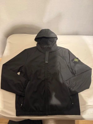 Svart vindjacka från Stone Island - Snygg svart vindjacka från Stone Island med ikonisk patch på vänster ärm. Jackan har huva, hel dragkedja framtill och två sidofickor med dragkedja. Tillverkad i ett lätt och slitstarkt material som passar perfekt för blåsiga dagar.
