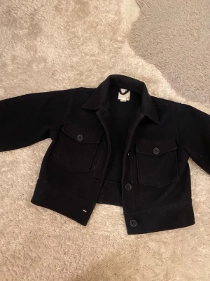 Svart overshirt med fickor, vår jacka  - Stilren svart overshirt med två stora bröstfickor och knappar framtill. Klassisk krage och raka ärmar. Perfekt att bära som lager över t-shirt eller hoodie. Materialet ser ut att vara mjukt och lite tjockare, troligen bomull eller en bomullsblandning.