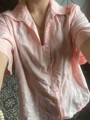 Ljusrosa skjorta med broderade detaljer - Säljer en ljusrosa skjorta med korta ärmar och broderade blommor. Skjortan har en avslappnad passform, klassisk krage och knäppning framtill. Materialet känns lätt och luftigt. 