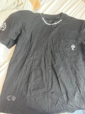 Svart Chrome Hearts t-shirt med tryck - Säljer en svart t-shirt från Chrome Hearts med vita tryckdetaljer på bröstfickan, ärmen och längs kragen. T-shirten har korta ärmar, rund halsringning och är tillverkad i mjuk bomull. Perfekt för dig som gillar streetwear och snygga detaljer.