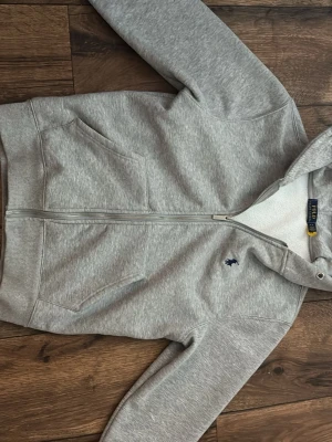 Grå hoodie från Polo Ralph Lauren - Snygg grå hoodie från Polo Ralph Lauren med dragkedja, två fickor fram och broderad logga på bröstet. Insidan är fodrad med mjuk teddy för extra komfort. Perfekt för chill dagar eller när du vill ha en clean look. Klassisk huva och ribbade muddar.