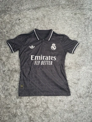Real Madrid svart matchtröja Adidas - Snygg svart Real Madrid matchtröja från Adidas med vita detaljer och klubbmärke på bröstet. Tröjan har klassisk krage, korta ärmar och tryckta loggor samt sponsortexten 'Emirates Fly Better' på framsidan. Perfekt för fotbollsträning eller att bära till vardags. För fler frågor/bilder skriv privat.