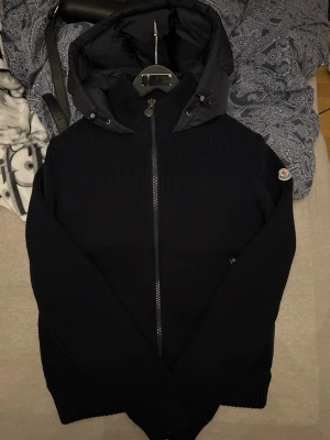 Svart pufferjacka från Moncler - Säljer en svart pufferjacka från Moncler med stickade ärmar och vadderad kropp. Jackan har en stor huva, dragkedja framtill och Moncler-logga på ärmen. Materialmix med dunfylld baksida och ribbade muddar för extra komfort och värme. Perfekt för kalla vinterdagar.