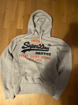Grå Superdry hoodie med tryck - Snygg grå hoodie från Superdry med stort tryck i marinblått och orange på bröstet. Klassisk känguruficka framtill, dragsko i huvan och ribbade muddar. Perfekt för en avslappnad streetstyle-look.