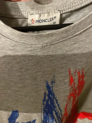 Grå Moncler t-shirt med tryck - Säljer en grå t-shirt från Moncler i bomull med ett stort grafiskt tryck i blått och rött på framsidan. Klassisk rund halsringning och korta ärmar. Perfekt för dig som vill ha en stilren men ändå unik look.