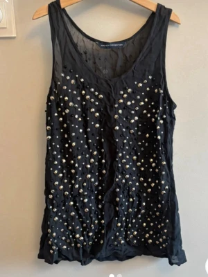 Svart pärlprydd linnetopp - Snygg svart linnetopp från French Collection, dekorerad med glittriga pärlor och nitar framtill. Toppen är ärmlös med rund halsringning och har en lätt transparent känsla. 