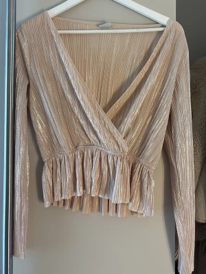 Champagnefärgad omlottopp med volang - Säljer en champagnefärgad omlottopp med lång ärm och glittrig finish. Toppen har djup v-ringning, plisserat tyg och volang nedtill för en extra snygg look, perfekt som en fest topp. Toppen är aldrig andvänd, pris kan diskuteras.