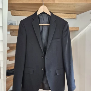 Svart klassisk kavaj herr - Stilren svart kavaj med två knappar framtill, klassisk passform och tre fickor. Fodrad insida och slits bak för extra komfort. Perfekt till skjorta och jeans eller chinos. Tidlös design som funkar till många looks.