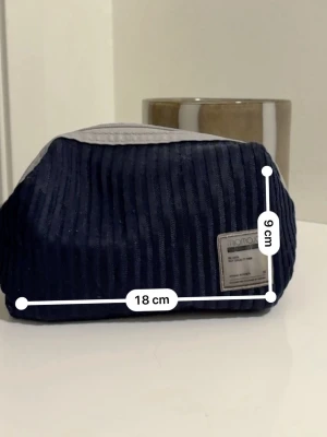 Smink väska - Säljer en mörkblå necessär från MOTTO i ribbad textil med grå detaljer och dragkedja upptill. Måtten är ca 18x9 cm. Perfekt för att organisera småsaker i väskan eller till resan. Snygg och enkel design med en liten etikett framtill.