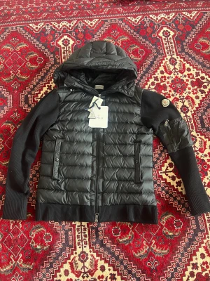 Svart  cardigan från Moncler - Snygg svart  cardigan från Moncler med quiltad front i glansigt material, stickade ärmar och rygg samt en vadderad huva. Jackan har dragkedja framtill, två fickor med tryckknapp och en patch med Moncler-logga på ärmen. Perfekt för kalla vinterdagar.