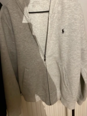 Grå zip hoodie från Polo Ralph Lauren - Snygg ljusgrå hoodie med dragkedja från Polo Ralph Lauren. Klassisk design med två fickor framtill och den ikoniska svarta loggan broderad på bröstet. Tillverkad i mjukt bomullsmaterial och har ribbade muddar vid ärmslut och nederkant.