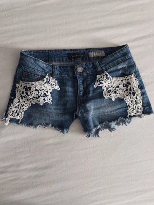 Blå jeansshorts med spets från Aeropostale - Snygga blå jeansshorts från Aeropostale med rå kant och vita spetsdetaljer på fickorna. Modellen är kort och har en klassisk femficksdesign. På bild tre ser de ljusare ut än i verkligheten.