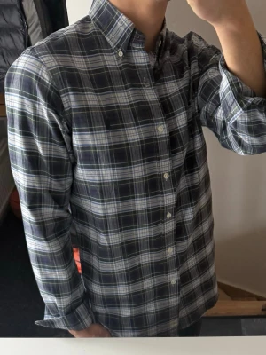 Mörkblå rutig skjorta med knappar - Snygg mörkblå rutig skjorta med vita och gröna detaljer. Skjortan har klassisk krage med knappar, långa ärmar och stängs med vita knappar framtill. Tillverkad i mjuk bomull, perfekt för en avslappnad men stilren look. Liten fläck där fram, därav priset 