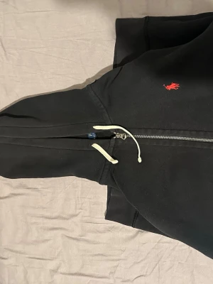 Svart hoodie från Polo Ralph Lauren - Klassisk svart hoodie från Polo Ralph Lauren med dragkedja framtill och vit dragsko i huvan. Ikonisk röd logga broderad på bröstet. Tillverkad i mjuk bomullsblandning som känns skön mot huden. Perfekt för en avslappnad och stilren look.