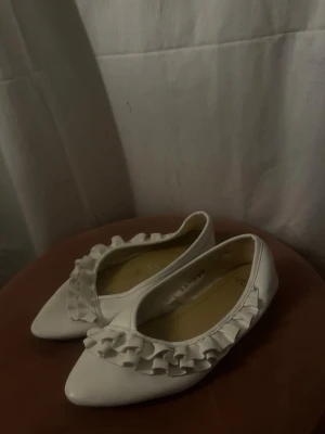 Vita ballerinaskor med volang - Säljer ett par vita ballerinaskor med spetsig tå och dekorativa volanger över foten. Skorna har en klassisk och feminin look, perfekta för dig som gillar detaljer. och materialet ser ut att vara konstläder.