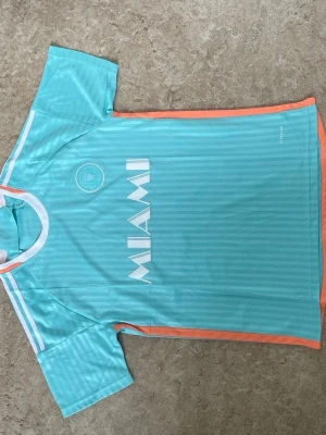 Turkos sporttröja MIAMI - Snygg turkos sporttröja med vita ränder och detaljer i orange. Framsidan har texten MIAMI och en logga på bröstet, baksidan har Royal Caribbean International-tryck. Tillverkad i lätt och ventilerande material, perfekt för träning eller sport.