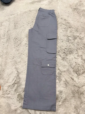 Grå cargobyxor med raka ben - Snygga grå cargobyxor med raka ben och flera praktiska fickor på sidorna, inklusive en med knappstängning. Byxorna har klassisk cargomodell med bälteshällor och är tillverkade i ett mjukt bomullsliknande material. Perfekta för en avslappnad och trendig stil.