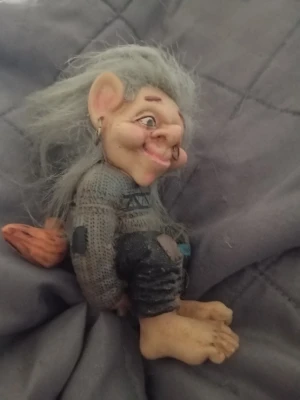 Trollfigur med grått hår - Säljer en trollfigur med långt, rufsigt grått hår och stora öron. Figuren har en stickad tröja i grått med mönster och mörka byxor. Trollet har stora fötter och sitter ner. Perfekt som dekoration eller samlarobjekt.