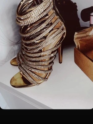 Guldiga sandaletter med strassdetaljer - Säljer ett par höga klackar i guld med öppna tår och många remmar täckta av glittrande strass. Skorna har en elegant siluett och klack, och är tillverkade i ett material som ser ut som metallicläder. Perfekta för dig som vill sticka ut och älskar bling.