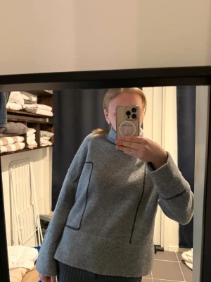 Grå stickad tröja från Zara - Snygg grå stickad tröja från Zara med hög krage och dekorativa svarta sömmar. Tröjan har en avslappnad passform, långa ärmar och ribbstickade muddar. Perfekt för lager på lager och enkel att matcha med olika stilar.