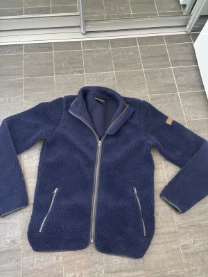 Mörkblå teddyjacka med dragkedja - Mysig mörkblå teddyjacka med hög krage och hel dragkedja framtill. Jackan har två snedställda fickor med dragkedja och en diskret patch på ena ärmen. Tillverkad i fluffigt teddy-material som håller dig varm under kyliga dagar.