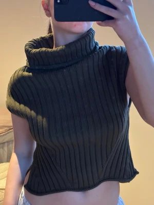 Stickad Topp - Säljer en olivgrön stickad topp med bred polokrage och ribbad struktur. Modellen är ärmlös och har en något kortare, boxig passform med vågig nederkant. Perfekt för lager på lager och trendiga outfits.🌸🩷☀️🌟