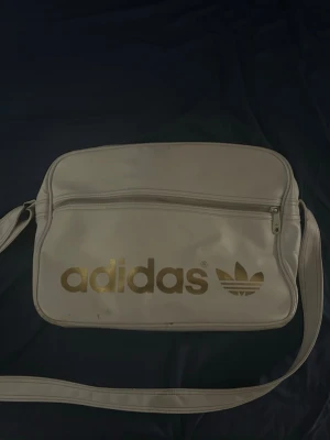 Vit axelväska från Adidas - Snygg vit axelväska från Adidas med guldigt logotryck och klassisk retrostil. Väskan har justerbar axelrem, stort huvudfack och en praktisk ytterficka med dragkedja. Tillverkad i konstläder som ger en clean och sportig look. Har lite sprickor på lädret vid dragkedjan som visas på bilden😊