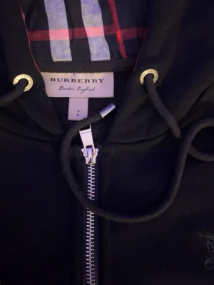 Svart Burberry hoodie med dragkedja - Svart hoodie från Burberry med klassiskt rutigt foder i huvan och insidan. Dragkedja framtill, snörning i huvan och guldfärgade detaljer vid snörhålen. Materialet är mjukt och bekvämt, perfekt för en cool och avslappnad look.