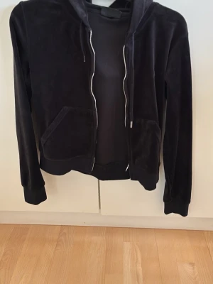 Svart velour hoodie från Juicy Couture - Mjuk och lyxig svart velour hoodie från Juicy Couture med dragkedja framtill, två fickor och broderad JC-logga på bröstet. Klassisk huva med snören och ribbade muddar vid ärmslut och nederkant. Perfekt för en chill och trendig look.
