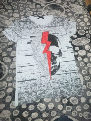 Grå t-shirt med dödskalle och blixt - Säljer en grå t-shirt med ett stort tryck av en dödskalle och en röd blixt framtill. T-shirten har korta ärmar och ett grafiskt mönster i svart och vitt. Blixten är dekorerad med små röda nitar för extra edge. Perfekt för dig som gillar statement-plagg.
