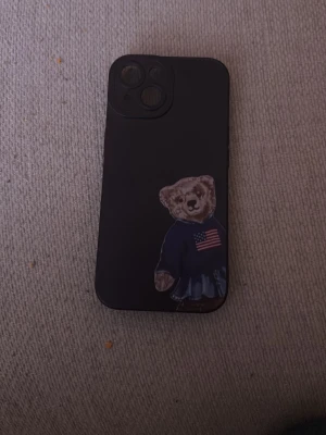 Björn mobilskal iPhone 15 - Snyggt svart mobilskal till iPhone 15 med en björn i blå tröja med amerikansk flagga. Skalet har öppningar för dubbla kameror och är gjort i ett flexibelt material som känns tåligt. Perfekt för dig som vill ha ett personligt och stilrent skydd till mobilen.