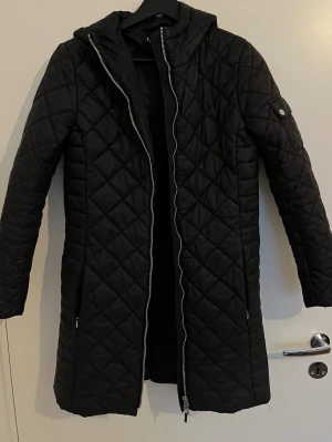 Svart lång dunjacka Lauren Ralph Lauren - Svart lång dunjacka från Lauren Ralph Lauren med quiltat mönster, huva och tvåvägsdragkedja. Jackan har fickor med dragkedja och en snygg knappdetalj på ärmen. Perfekt för kalla vinterdagar och ger en clean och stilren vibe.