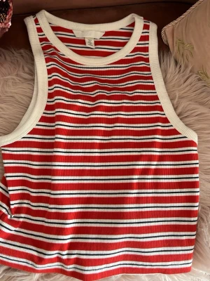 Randig ribbad linnetopp röd/vit - Snygg ribbad linnetopp med röda, vita och svarta ränder. Modellen är ärmlös med breda axelband och rund halsringning. Perfekt för varma dagar och enkel att styla med jeans eller kjol.