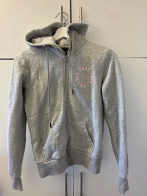 Grå zip-hoodie från Peak Performance - Snygg grå hoodie med dragkedja från Peak Performance. Har huva med dragsko, fickor framtill och rosa broderad logga på bröstet. Tillverkad i mjukt sweatshirtmaterial som är perfekt för chill eller träning.