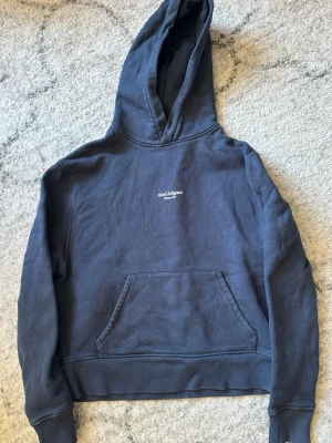 Mörkblå croppad hoodie från Axel Arigato dam - Snygg mörkblå hoodie från Axel Arigato med liten vit logga på bröstet. Klassisk känguruficka framtill och ribbade muddar vid ärmar och nederkant. Tillverkad i mjuk bomull för skön känsla och avslappnad passform. Perfekt till streetwear-looken. Hoodien är i storlek S för dam, croppad modell. Använd men i mycket bra skick