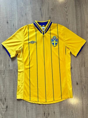 Svensk landslagströja Umbro gul retro - Gul svensk retro landslagströja från Umbro från säsongen 2012/2013 med blå tunna ränder, blå detaljer vid krage och ärmslut samt broderad svensk landslagssköld på bröstet. Tillverkad i lätt och ventilerande material, perfekt för VM i sommar🟡🔵