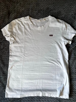 Vit t-shirt från Levi's - Klassisk vit t-shirt från Levi's med rund halsringning och liten röd logotyp på bröstet. T-shirten är enkel och stilren, perfekt till jeans eller shorts. Tillverkad i mjuk bomull och har korta ärmar.