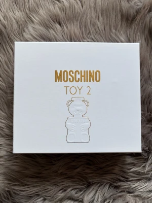 Moschino Toy 2 presentbox - Lyxig presentbox från Moschino med Toy 2 parfym och matchande body lotion och shower gel. Flaskan har en unik björnform i frostat glas med gulddetaljer. Tuben är vit med guldtext och björnmotiv. Perfekt för dig som gillar lekfull och trendig design. ( Helt ny )