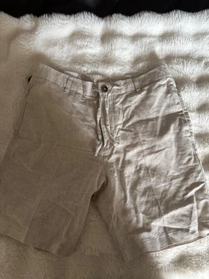 Beiga linneshorts från Selected Homme - Snygga beiga linneshorts från Selected Homme med knäppning fram, dragsko i midjan och bälteshällor. Shortsen har två bakfickor med knapp och är tillverkade i ett luftigt linnematerial – perfekt för varma dagar.
