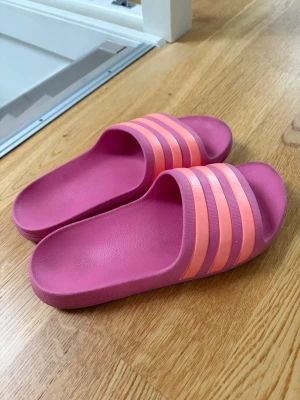 Rosa slides med ränder från Adidas - Säljer ett par snygga rosa tofflor från Adidas med tre ljusrosa ränder över foten. Tofflorna är gjorda i ett mjukt och bekvämt syntetmaterial, perfekta att glida i hemma eller på stranden. Klassisk slip-on-modell med sportig vibe. Något smutsiga men fint skick!💕Kan rengöras innan säljning💕