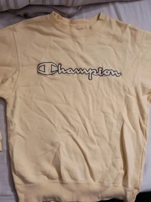 Gul sweatshirt från Champion - Snygg gul sweatshirt från Champion med stort broderat logotyptryck över bröstet. Tröjan har rund halsringning och ribbade muddar vid ärmslut och nederkant. Perfekt för en avslappnad och sportig stil. Osäker på passform men antingen unisex eller kvinna. 