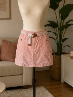 Rosa kortkjol med bälte - Söt rosa kortkjol med dekorativt bälte i vitt och rosa med brunt spänne. Kjolen har två knappar framtill och en avslappnad passform, perfekt för en trendig vardagslook.