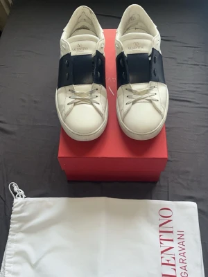 Valentino open - Snygga vita sneakers från Valentino Garavani med bred svart läderdetalj över vristen och ikoniska nitar på hälen. Skorna har klassisk låg profil och är tillverkade i läder med vita snören. Perfekta för en trendig och stilren look.
