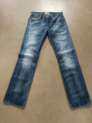 Blå raka jeans från Levi's - Säljer ett par klassiska blå jeans från Levi's med rak passform och fem fickor. Jeansen har en snygg tvättad look med slitningar och kontrastsömmar. Tillverkade i slitstark denim med Levi's ikoniska läderpatch bak i midjan. 