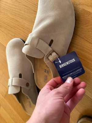 Beige Birkenstock Boston tofflor - Snygga beige Birkenstock Boston tofflor i mocka med justerbart spänne och klassisk korksula. Yttersulan är svart och har ett mönstrat grepp. Perfekta för en chill och stilren look. HELT OANVÄNDA SKULLE KÖPT STRL 45 