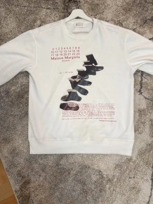 Maison Margiela vit sweatshirt med tryck - Säljer en clean vit sweatshirt från Maison Margiela med unikt grafiskt tryck framtill och röd text. Tröjan har rund halsringning och ribbade muddar. Perfekt för dig som gillar statement-plagg och vill sticka ut med en exklusiv design.