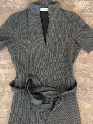Grå jumpsuit med bälte från Mango - Stilren grå jumpsuit från Mango med korta ärmar, markerad krage och knytbälte i midjan. Vida ben och diskret mönster ger en modern look. Perfekt för dig som gillar minimalistisk stil och vill ha något bekvämt men snyggt.