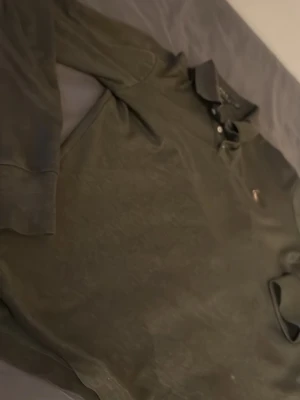 Mörkgrön långärmad polotopp - Snygg mörkgrön långärmad polotopp med klassisk krage och knappar framtill. Liten broderad logga på bröstet. Perfekt för en stilren och avslappnad look. Liten fläck, går å tvättad bort! Perfekt inför sommaren!