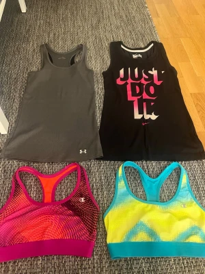 Träningslinnen & sport-bh från Nike, Under Armour & Champion - Fyra sportiga plagg: ett grått linne från Under Armour, ett svart linne från Nike med 'Just Do It'-tryck i rosa och vitt, samt två sport-bh:ar från Champion i färgglada mönster – en i rosa/röd och en i blå/gul. Perfekta för träning och gym.  Sportbh i storlek M. Kan köpa alla eller bara någon av de så skriv!